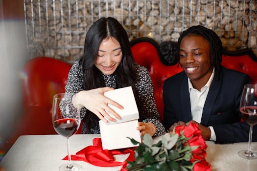 Un couple souriant ouvre un cadeau sur une table décorée de roses et de verres de vin rouge.